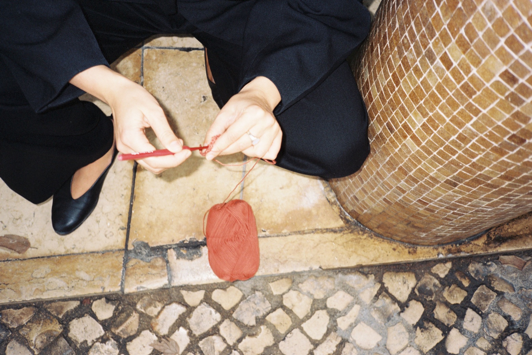Hands crocheting with red yarn while sitting beside a tiled column